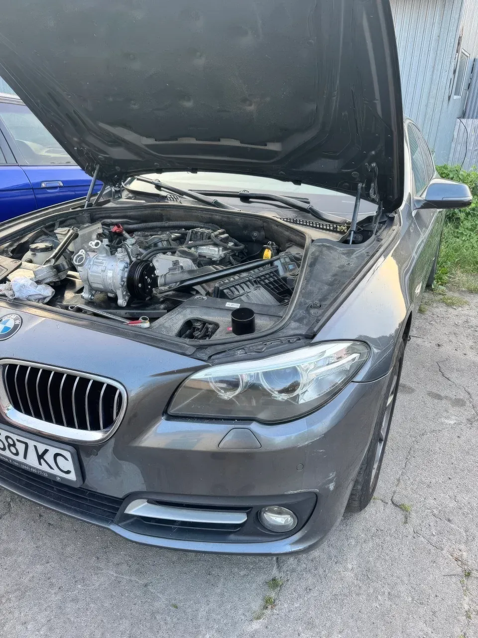 На відкритому майданчику показано сірий BMW із піднятим капотом; у моторному відсіку видно компресор кондиціонера, ремінний привід та робоче приладдя. Людей у к